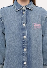 Gaiši zila džinsu krekls ar pogu aizdari, sudraba pogām un rozā logo uzsieti "MOSCHINO JEANS" kreisajā pusē.