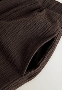 Pantalones de tela texturizada de color marrón oscuro con cintura elástica y un bolsillo lateral profundo, mostrados sobre una superficie clara.