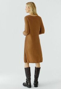 Braun gestricktes Kleid mit langen Ärmeln, rundem Ausschnitt und seitlichen Schlitzen; kombiniert mit knielangen schwarzen Lederstiefeln. Weiches, strukturiertes Material.