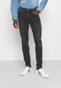 Vaqueros negros ajustados con agujeros desgastados en las rodillas, combinados con una camisa de mezclilla azul clara y zapatillas blancas con detalles rojos.