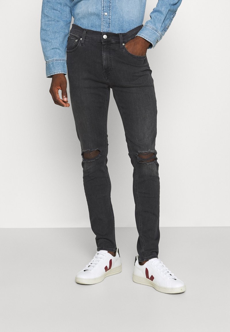 Vaqueros negros ajustados con agujeros desgastados en las rodillas, combinados con una camisa de mezclilla azul clara y zapatillas blancas con detalles rojos.