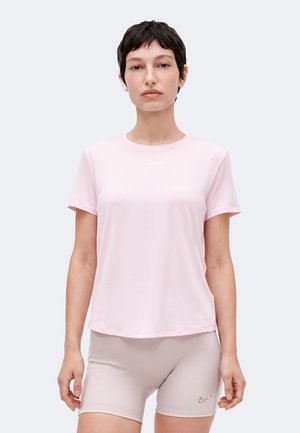 Femme aux cheveux courts et foncés portant un t-shirt rose clair à manches courtes et un short de cyclisme beige, debout devant un fond uni.