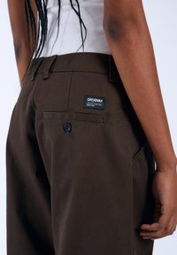 Bruine broek van een duurzame stof, met een achterzak met een zwart label waarop "DRDENIM" staat en een knoopsluiting. Glad textuur.