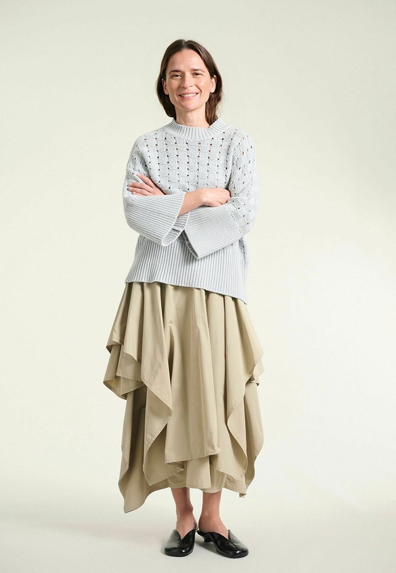 Femme souriante debout, les bras croisés, portant un pull en maille gris clair, une jupe beige à plusieurs couches et des chaussures noires sans lacets sur un fond clair.