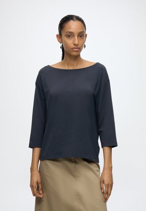 Vrouw met strak naar achter gekamd haar draagt een marineblauwe top met driekwartsmouwen en een beige rok, staand tegen een effen lichte achtergrond.