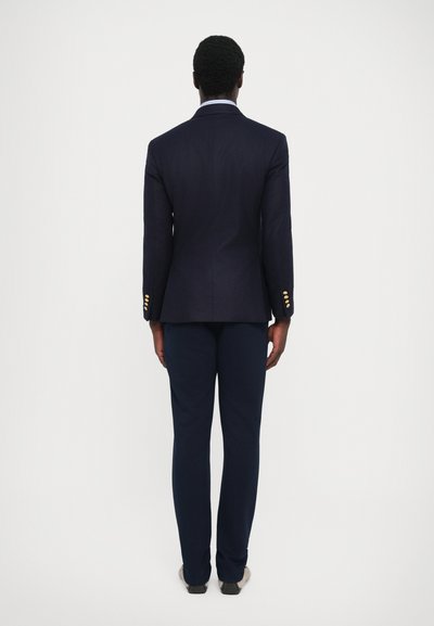Veste de costume bleu marine avec des accents de boutons dorés, coupe ajustée, associée à un pantalon assorti, affichant une texture lisse et un design moderne.