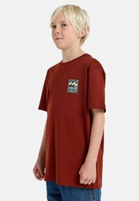 Camiseta de algodón color óxido con mangas cortas, que presenta un parche gráfico cuadrado en verde y azul, sobre un fondo liso.