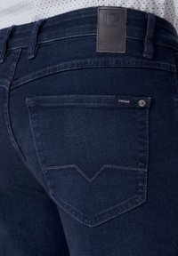 Nahaufnahme einer dunkelblauen Jeans-Hosentasche mit sichtbaren Nähten und einem Leder-Brandlabel am Bund.