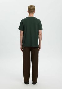 Camiseta de algodón verde oscuro con cuello redondo estándar, mangas cortas y un corte relajado, combinada con pantalones marrones de corte holgado.