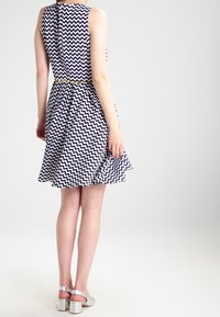 Robe sans manches avec un motif en zigzag bleu marine et blanc, jupe évasée, taille cintrée et ceinture jaune. Des sandales à talons argentées complètent le look.