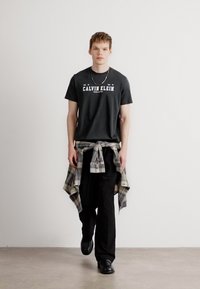Mustmust särk, millel on "CALVIN KLEIN" logo, särgiga, mis on seotud talje ümber, laiad mustad püksid ja mustad kingad, neutraalse tausta ees.