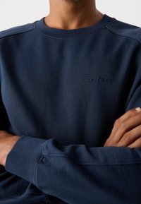 Uomo che indossa una felpa a maniche lunghe di colore blu navy con le braccia incrociate, con il logo ricamato "Marc O'Polo" sul petto.
