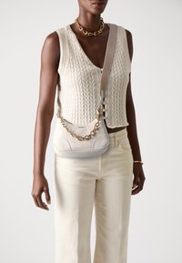 Grey leather shoulder bag with embossed floral pattern, gold chain accent, and adjustable strap. Worn with a knitted sleeveless top and cream pants.