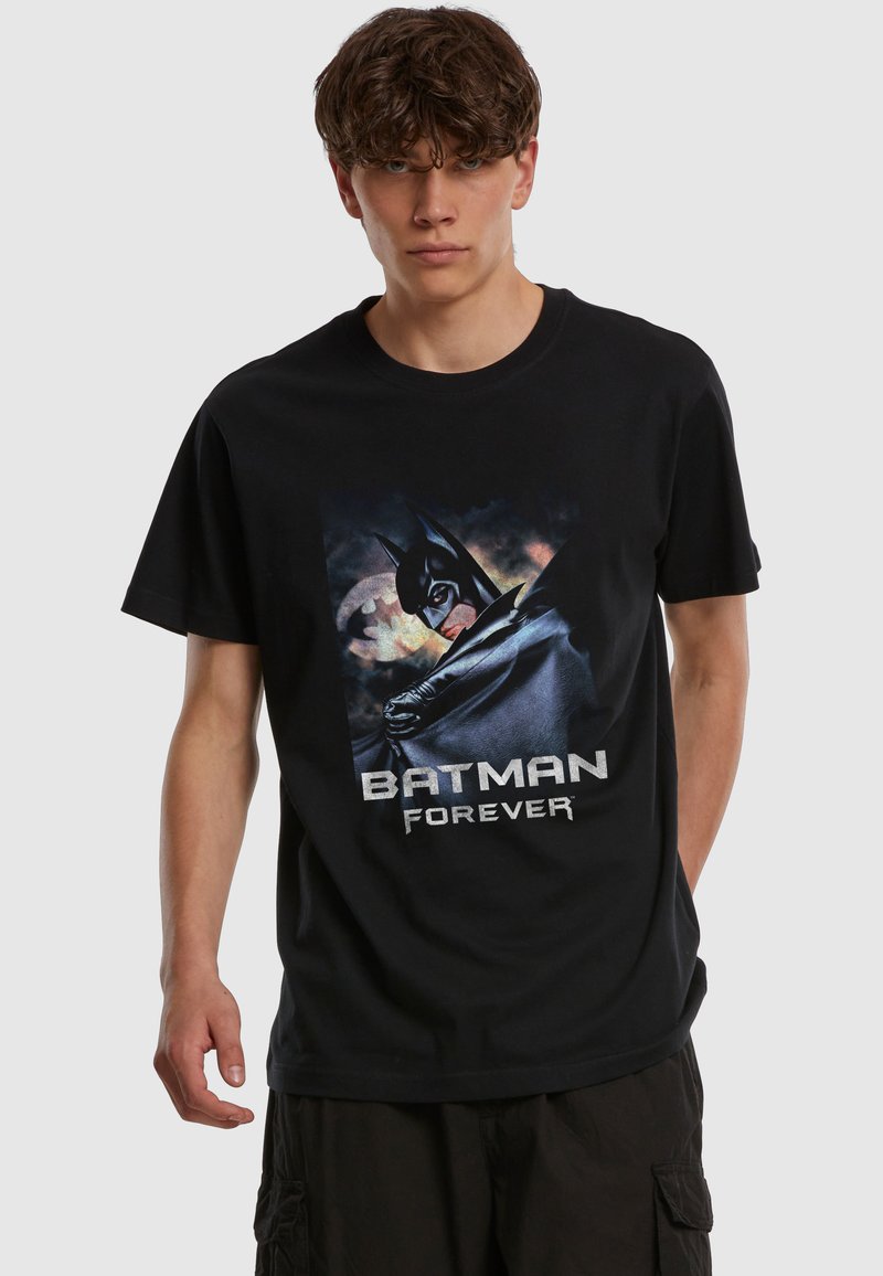 Jeune homme portant un t-shirt noir "Batman Forever" avec un graphisme Batman et un pantalon cargo sombre sur un fond uni.