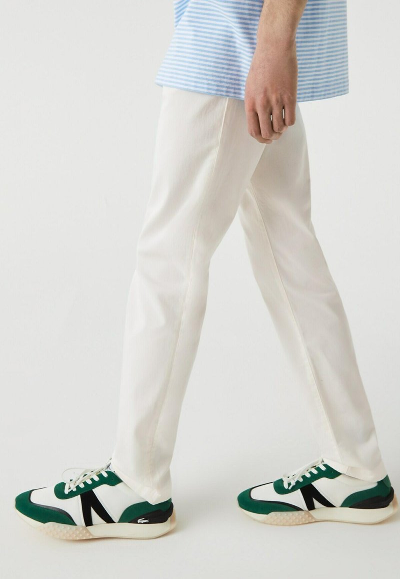 Persona che cammina con pantaloni bianchi e sneakers bianchi, verdi e neri su uno sfondo bianco semplice.