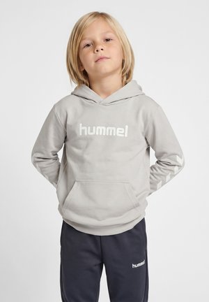Małe dziecko o blond włosach, ubrane w jasnoszarą bluzę z kapturem Hummel i ciemne spodnie Hummel, stoi z rękami za plecami na jednolitym tle.