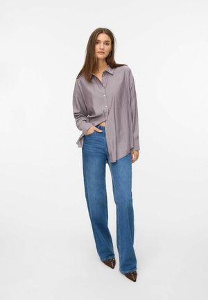 Chemise rayée boutonnée en violet clair et blanc, assortie à un jean évasé bleu. La chemise a une coupe décontractée et des manches retroussées.