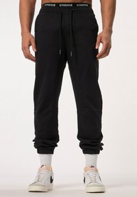 Pantalones jogger negros con puños elásticos y cintura con cordón, que presentan bolsillos laterales y detalles de costura sutiles. Combinados con zapatillas blancas.