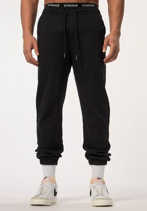 Pantalones jogger negros con puños elásticos y cintura con cordón, que presentan bolsillos laterales y detalles de costura sutiles. Combinados con zapatillas blancas.