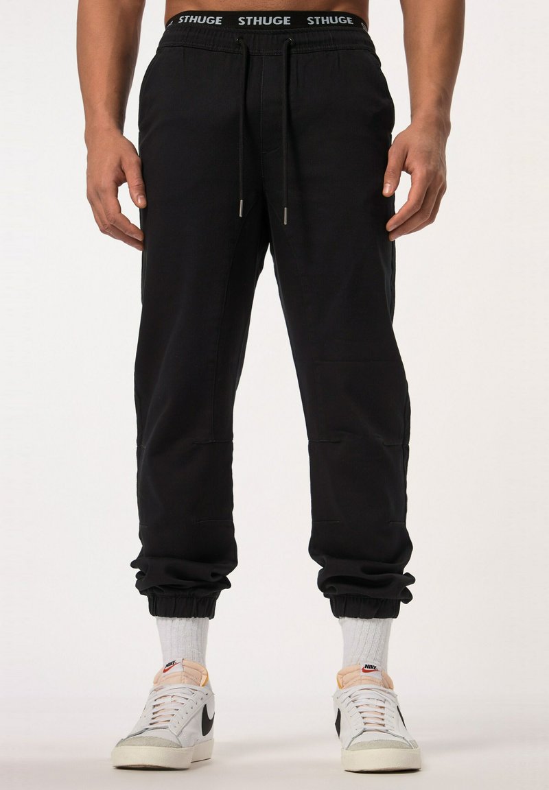 Sorte joggingbukser med elastiske cuffs og snor i taljen, der har sidelommer og subtile stiksyninger. Sammen med hvide sneakers.