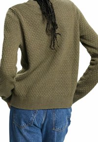 Personne portant un pull en maille texturée vert olive et un jean en denim bleu, vue de dos avec les mains sur les hanches.