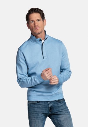 Man in een blauw pullover met kwart rits en spijkerbroek, die zijn mouw verstelt, staand tegen een effen lichte achtergrond.