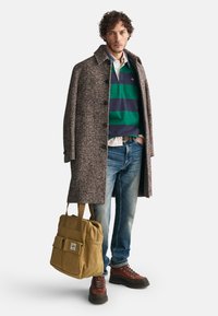 Brūns zivtiņu raksta mētelis pār zaļi un tumši zilu svītrainu džemperi, zilas džinsa bikses, brūni pārgājienu zābaki un tan auduma soma ar utilitārajiem kabatām.