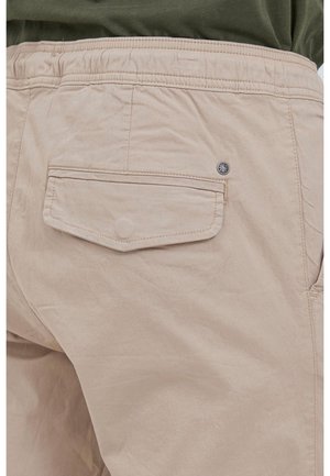 Pantalon beige clair avec taille élastique et poche arrière à rabat sécurisée par un bouton caché et un petit détail logo métallique.