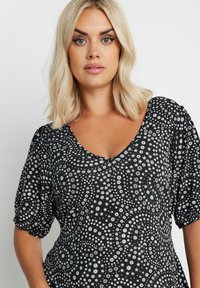 Mustapohjainen kukkatoppi lyhyillä puffhihoilla, jossa on valkoisia abstrakteja kukka- ja pisarakuoseja, V-pääntie ja pehmeä, kevyt kangas.