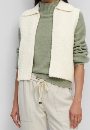 Weißer, flauschiger Zip-Up-Weste über einem salbeigrünen Strickpullover und cremefarbenen Hosen mit Kordelzug, die den Oberkörper und die Arme entspannt an den Seiten zeigt.
