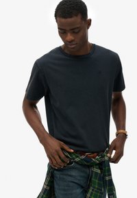 Marineblå bomulds t-shirt med rund hals, korte ærmer og et lille logo på brystet; modellen har denim jeans og en grønternet skjorte bundet om livet.
