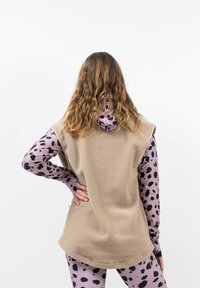 Beige Fleece-Weste mit abgerundetem Saum, getragen über einem pinken Langarmoberteil mit schwarzen Punkten. Die Rückseite hat keine sichtbaren Verschlüsse oder Akzente.