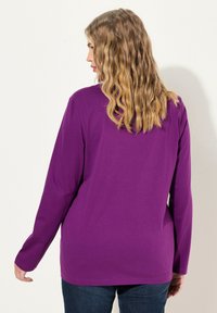 Fialové tričko s dlouhým rukávem, volného střihu, hladkého textilu a kulatým výstřihem, v kombinaci s tmavě modrými džíny, viděné zezadu.