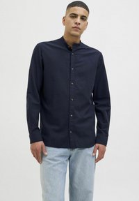 Camicia a maniche lunghe blu navy con colletto alla cinese, caratterizzata da chiusura con bottoni e una texture liscia, abbinata a jeans blu chiaro.