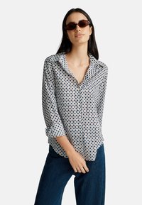 Donna con lunghi capelli scuri che indossa occhiali da sole tartarugati, una camicia a maniche lunghe con motivo e maniche arrotolate, e jeans blu scuro.