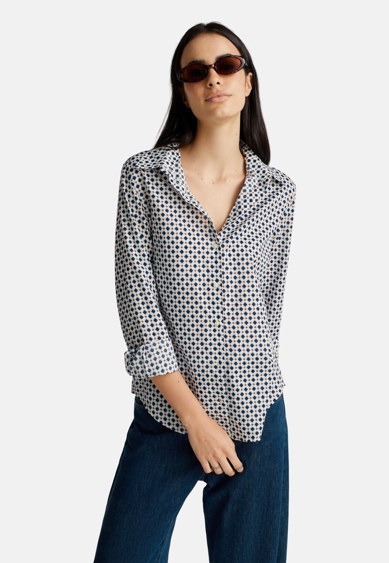 Donna con lunghi capelli scuri che indossa occhiali da sole tartarugati, una camicia a maniche lunghe con motivo e maniche arrotolate, e jeans blu scuro.