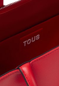 Bolso de mano rojo con textura lisa, que presenta un logotipo plateado de "TOUS". Incluye un cierre de cremallera y un borde negro a contraste.