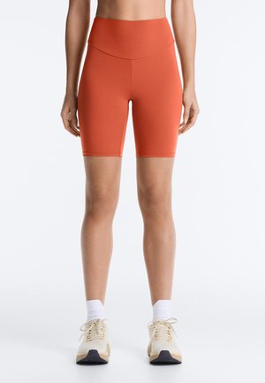 Donna che indossa pantaloncini da ciclista arancioni a vita alta, calzini bianchi alla caviglia e scarpe sportive beige, in piedi su uno sfondo bianco semplice.