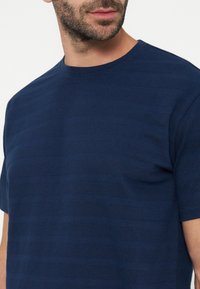 Homme portant un t-shirt texturé bleu marine à manches courtes et col rond, montrant le haut du torse et une partie du visage avec barbe.