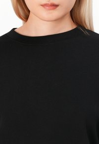 Sweatshirt noir à col rond fabriqué en tissu doux et lisse. Il présente un col en rib et un design minimaliste sans motifs visibles.