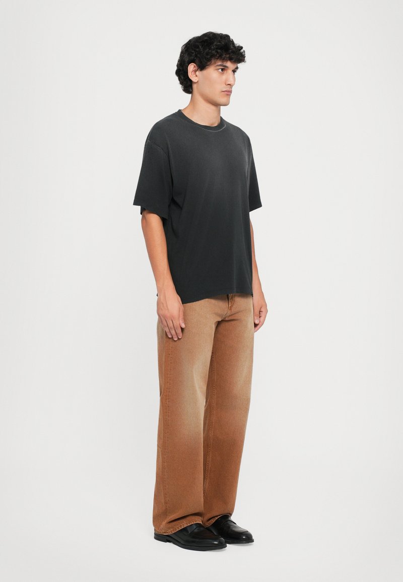 Black cotton t-shirt with loose fit, paired with light brown wide-leg jeans and black shoes, standing against a plain white background.