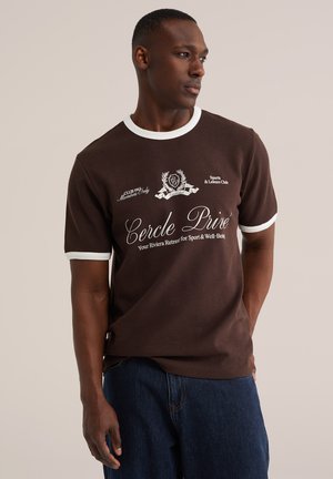 Mężczyzna noszący brązowy t-shirt z białą lamówką i nadrukowanym tekstem, w połączeniu z niebieskimi dżinsami, stoi na tle jednolitego beżowego tła.