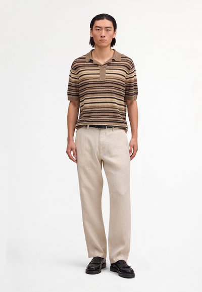 Mann trägt ein kurzärmeliges gestreiftes braunes Poloshirt, beige Hose, schwarzen Gürtel und schwarze Loafers, steht vor einem weißen Hintergrund.
