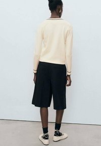 Maglione a maniche lunghe crema con dettaglio nero abbinato a pantaloni corti neri larghi e scarpe chiare. Tessuto liscia, vestibilità rilassata e design minimalista.
