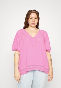 Blusa rosa com decote em V, mangas curtas e detalhamentos em renda ao longo do decote e da bainha. Combinada com jeans azul claro.