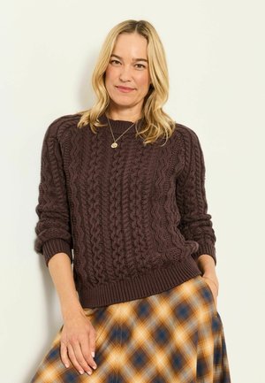 Kobieta o blond włosach, ubrana w ciemnobrązowy sweter w warkocze oraz żółto-niebieską spódnicę w kratę, stojąca na jasnym tle.