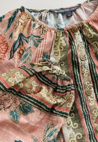 Bluse im Boho-Stil mit floralen und geometrischen Mustern in Pink, Grün und Beige, ausgestattet mit Holzknöpfen und gerafften, elastischen Ärmeln.