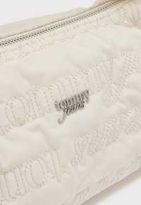 Bolsa de tela crema con patrón acolchado de texto "tommy jeans" y logo de metal plateado "tommy jeans", con una cremallera en la parte superior.