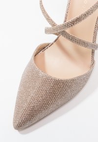 Chaussure à talon aiguille à bout pointu en tissu métallisé rose doré avec une surface texturée, dotée de brides entrecroisées au niveau de la cheville.