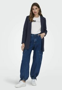 Blazer bleumarin peste un tricou alb cu grafică, asortat cu pantaloni cargo lejeri din denim albastru și adidași albi. Are manșete elastice și detalii cu buzunare.